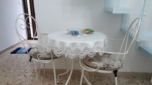 Un petit coin repas avec une table ronde recouverte d’une nappe en dentelle blanche, deux chaises en métal et une petite table sur laquelle sont disposés des aliments.