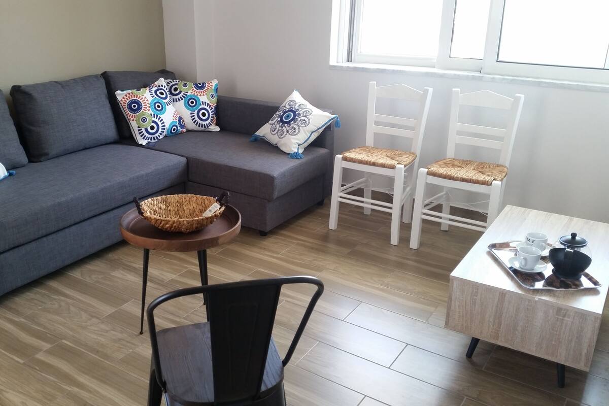Ein modernes Wohnzimmer mit einem grauen Sofa, gemusterten Kissen, einem gewebten Korb auf einem kleinen Tisch, zwei weißen Stühlen mit gewebten Sitzflächen und einem Couchtisch mit einer Kanne und Tassen.