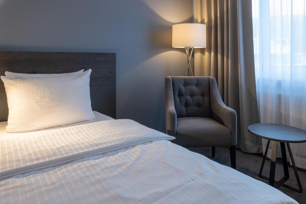 A hotel room with a bed, a chair, a small table, and a lamp.