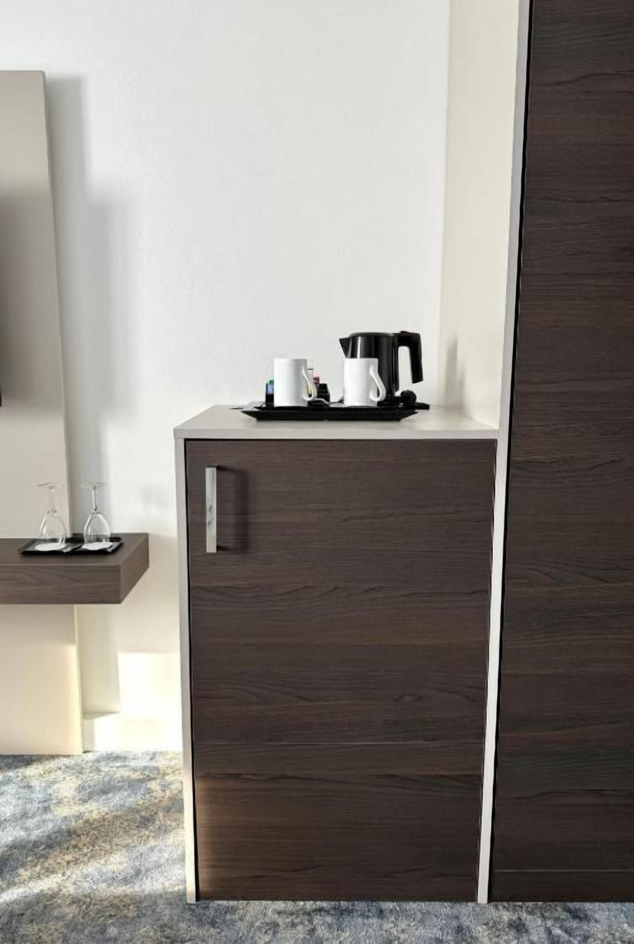 Een donkere houten kast met een koffiezetapparaat en kopjes bovenop, een klein plankje met glazen, en een spiegel aan de linkerkant.
