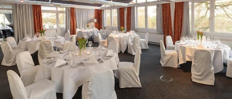 A banquet hall with round tables set for dining, white chairs, and large windows with curtains.