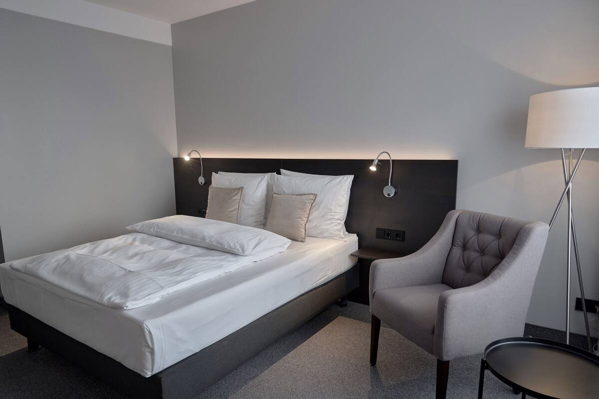 Ein modernes Hotelzimmer mit einem ordentlich bezogenen Bett, einem Sessel und einer Lampe.