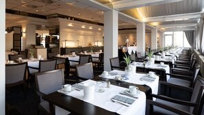 Un'area pranzo moderna con diversi tavoli apparecchiati, una cucina sullo sfondo e vista sulla sala da pranzo attraverso ampie finestre.
