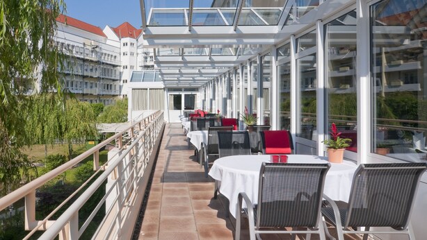 Un'area pranzo all'aperto illuminata dal sole, con tavoli e sedie, una passerella vetrata e vista sugli edifici sullo sfondo.