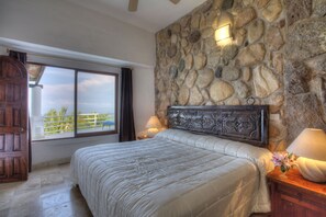 A bedroom with a stone wall, a large bed, a wooden door, and a view of the outdoors.