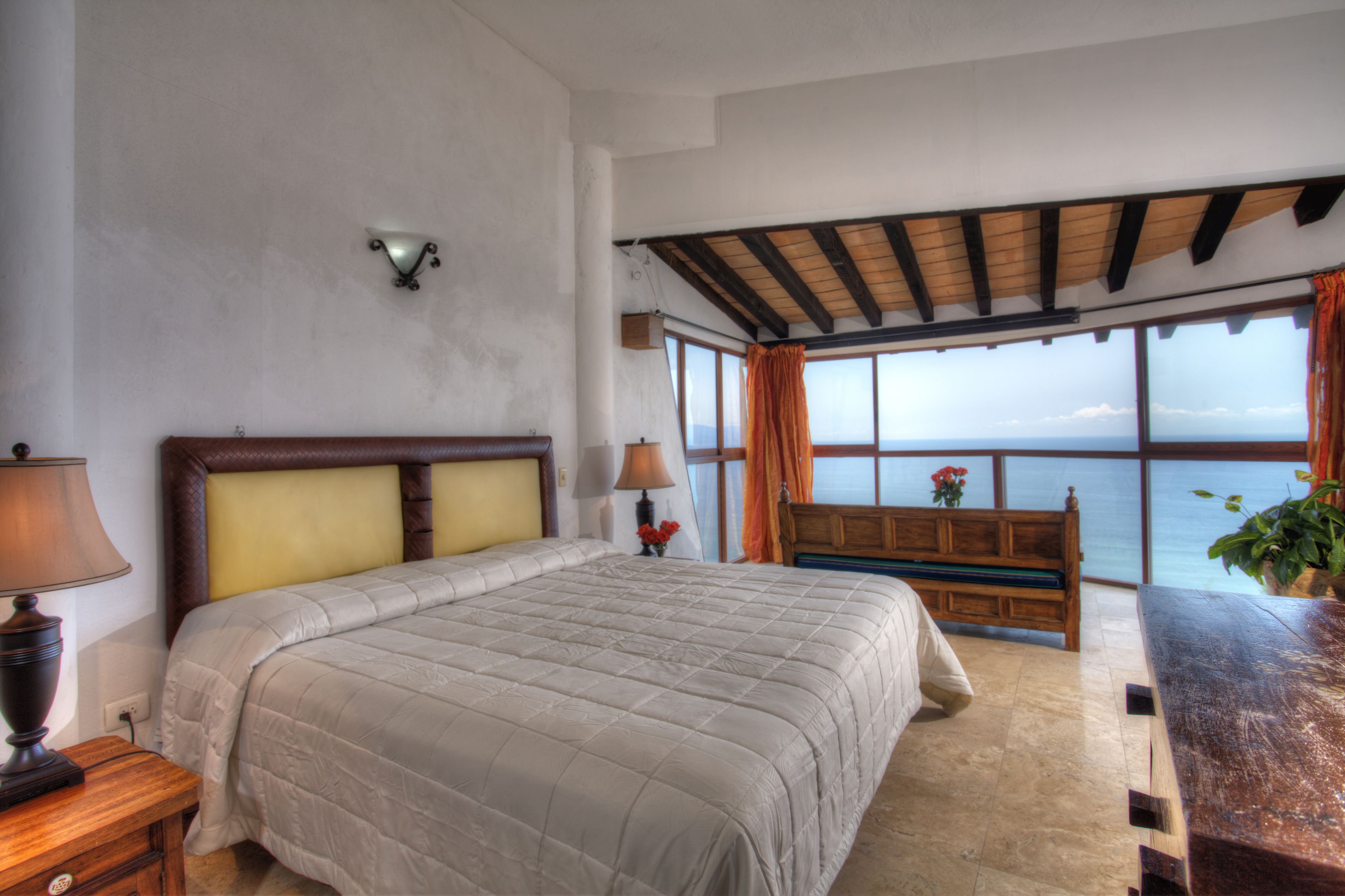 Een slaapkamer met een groot bed, een houten bankje, een tafel en uitzicht op zee.