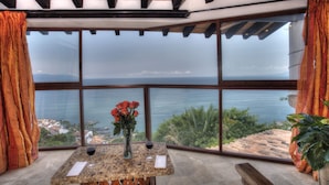 Una habitación con un ventanal amplio que da al mar, una mesa con un florero y una copa de vino tinto.