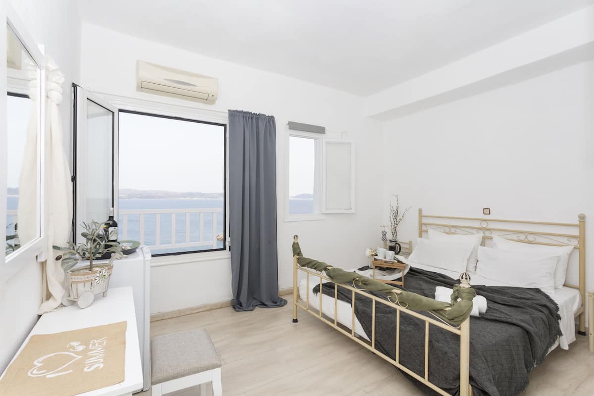A bedroom with a bed, a bedside table, a window with curtains, a potted plant, and a view of the sea.