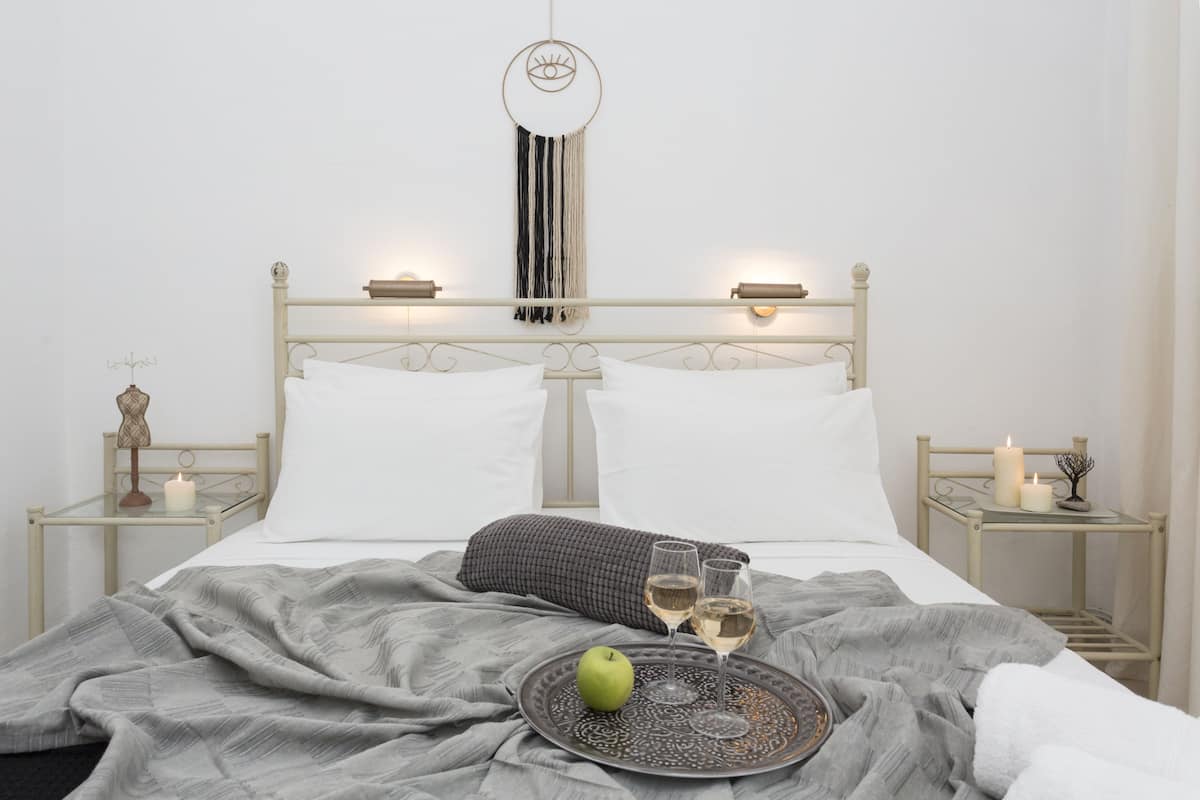 A neatly made bed with white linens, a gray blanket, and a tray with a green apple and two glasses of wine.