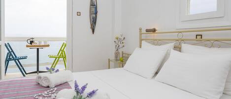 Una cama bien hecha con sábanas blancas, un tapete de baño a rayas y una decoración con una estrella de mar. Hay una mesita con una taza y un platillo, y una silla con asiento verde.