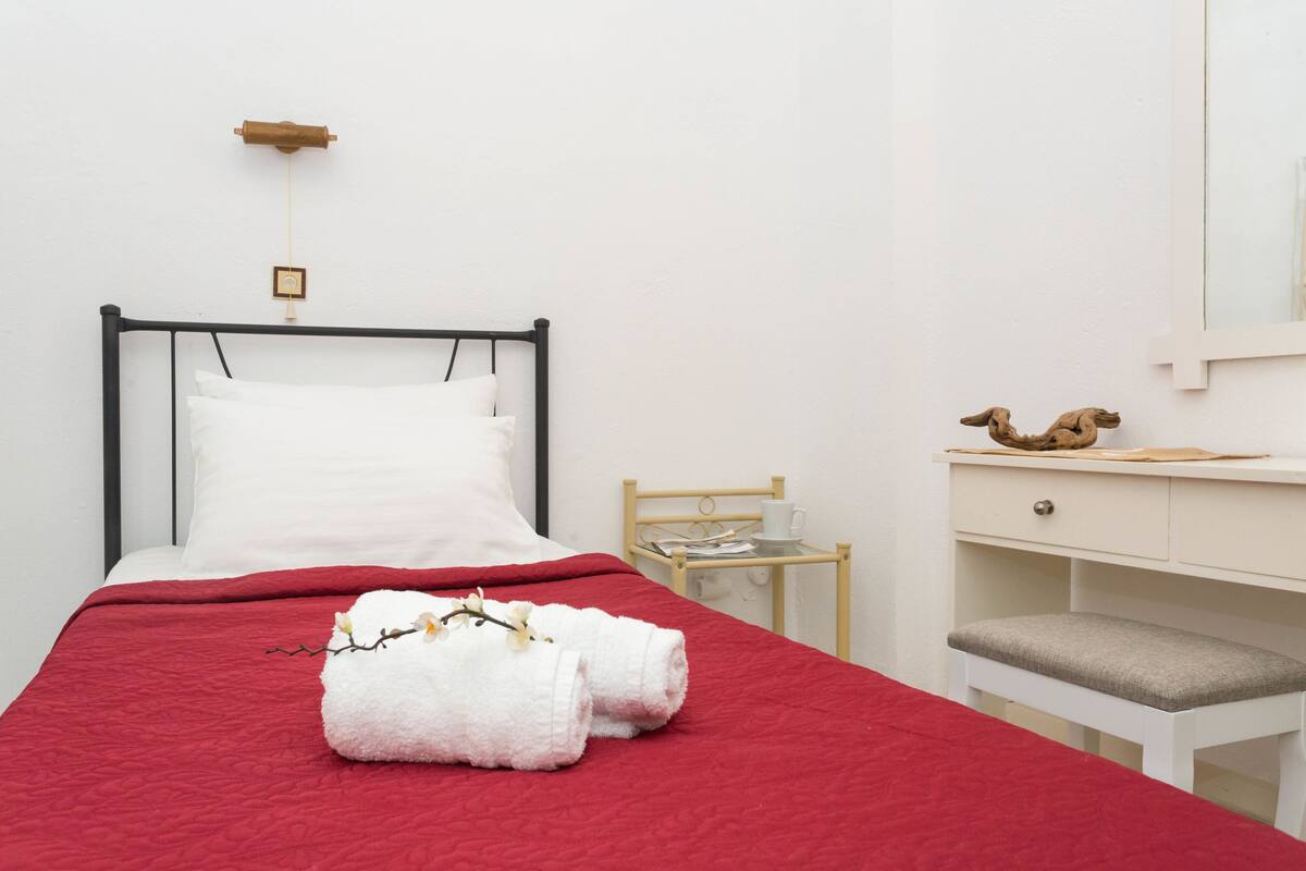 A neatly made bed with a red bedspread and white pillows, a wooden nightstand with a cup and saucer, and a white desk with a bench.