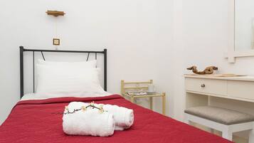 Una cama con una manta roja, almohadas blancas y una ramita con flores blancas. Hay una silla de madera y un escritorio blanco con una taza y un platillo.