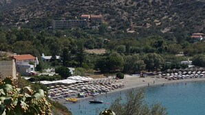 A coastal town with a beach lined with umbrellas and sun loungers, surrounded by hills and buildings. - Galini Mare (Agios Vasileios)