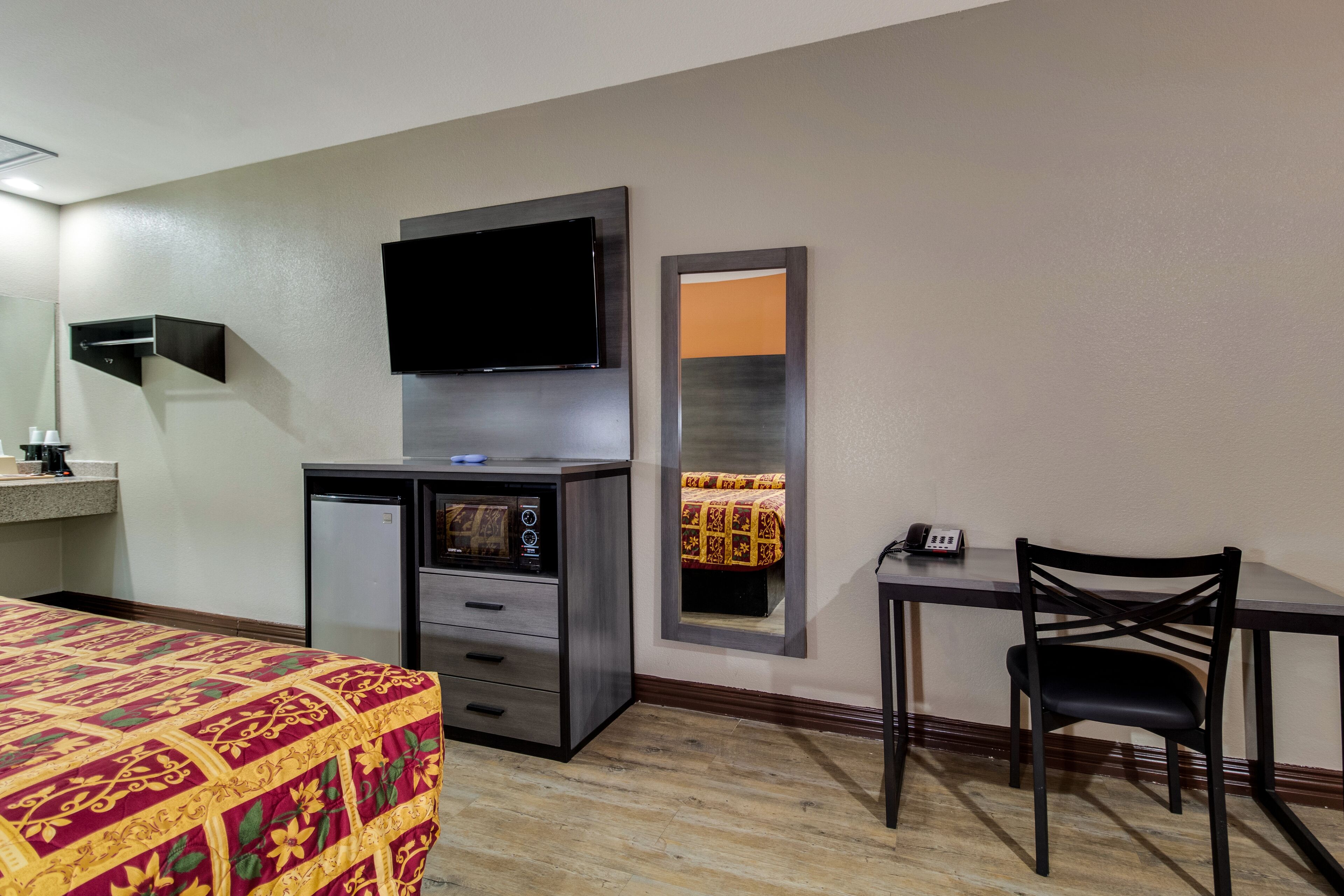 A hotel room with a bed, a desk with a chair, a television mounted on the wall, a small refrigerator, and a mirror.