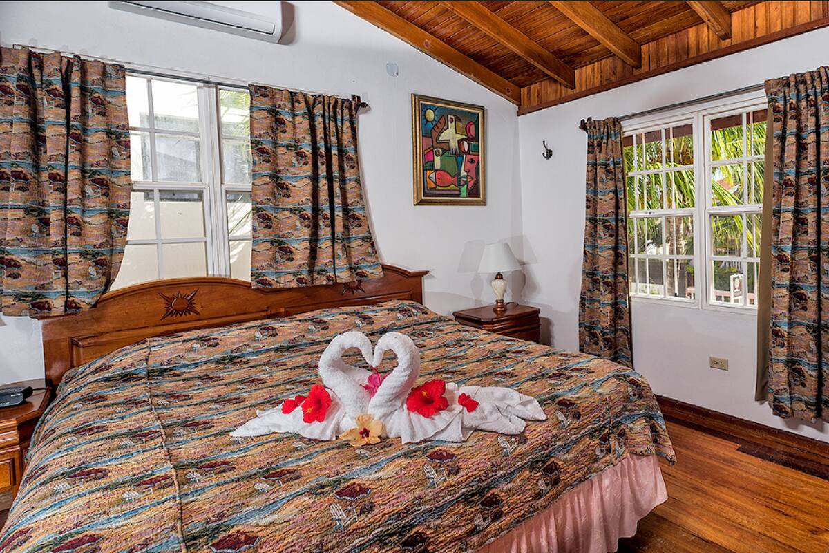 Quarto de hotel com teto de madeira, cama com colcha florida, duas janelas com cortinas e um quadro na parede.