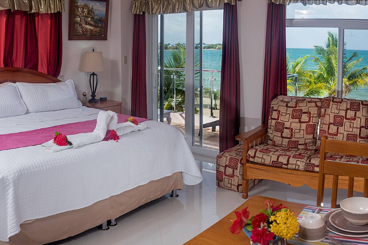 Quarto de hotel com cama, poltrona, mesa com um vaso de flores, luminária e vista para o oceano.
