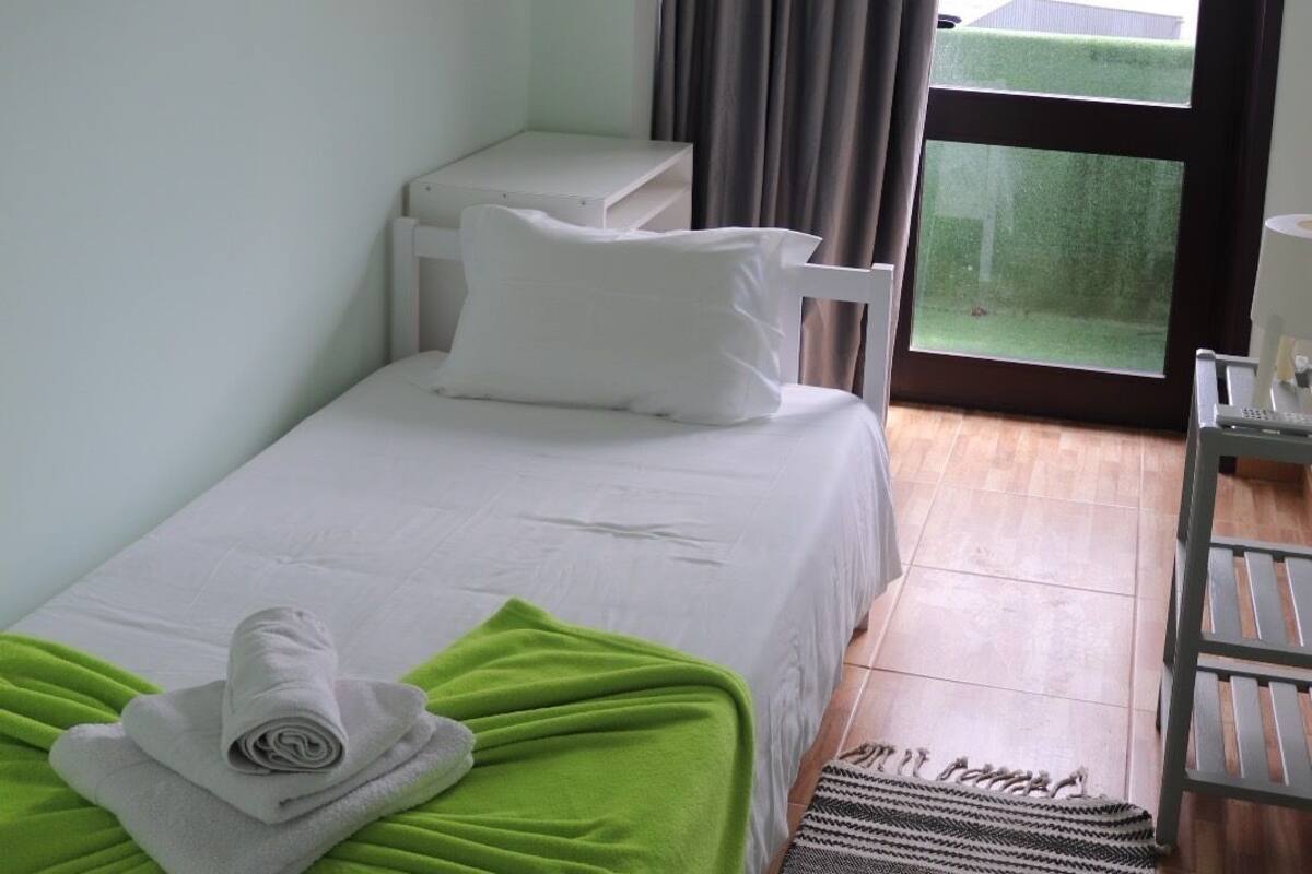 A single bed with a green blanket and rolled towels, a white bedside table, a small white chair, and a view of a green lawn through a large window.