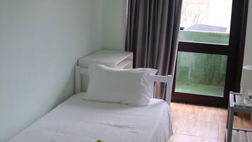 Un lit simple avec une couverture verte et des serviettes enroulées, une table de chevet blanche, une petite chaise blanche et une vue sur une pelouse verdoyante par une grande fenêtre.