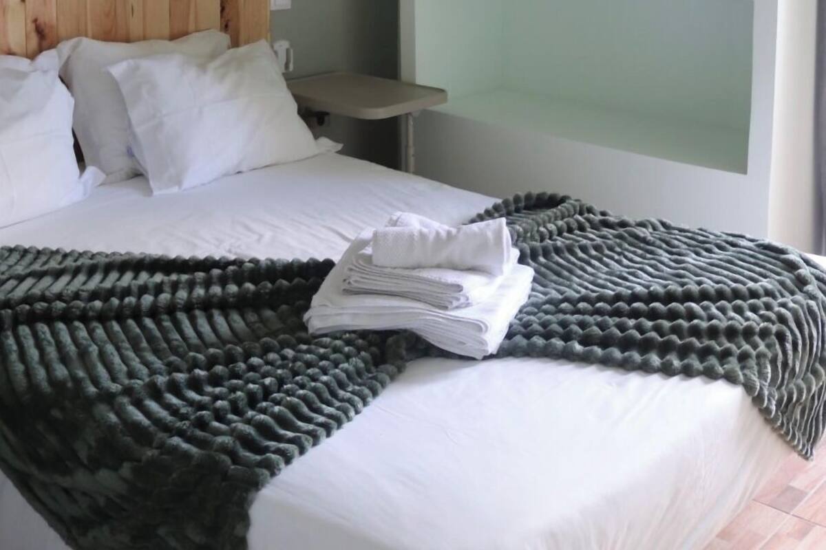 A bedroom with a wooden headboard, a bed with white linens, a dark green textured blanket, and a wall-mounted shelf with a hanging rack.