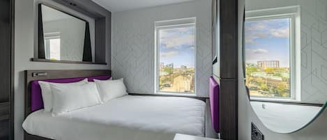 Habitación de hotel moderna con una cama grande, un tocador con espejo y dos ventanas con vista a la ciudad.