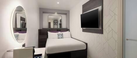A modern hotel room with a bed, a TV mounted on the wall, a bedside table with a lamp, and a mirror.