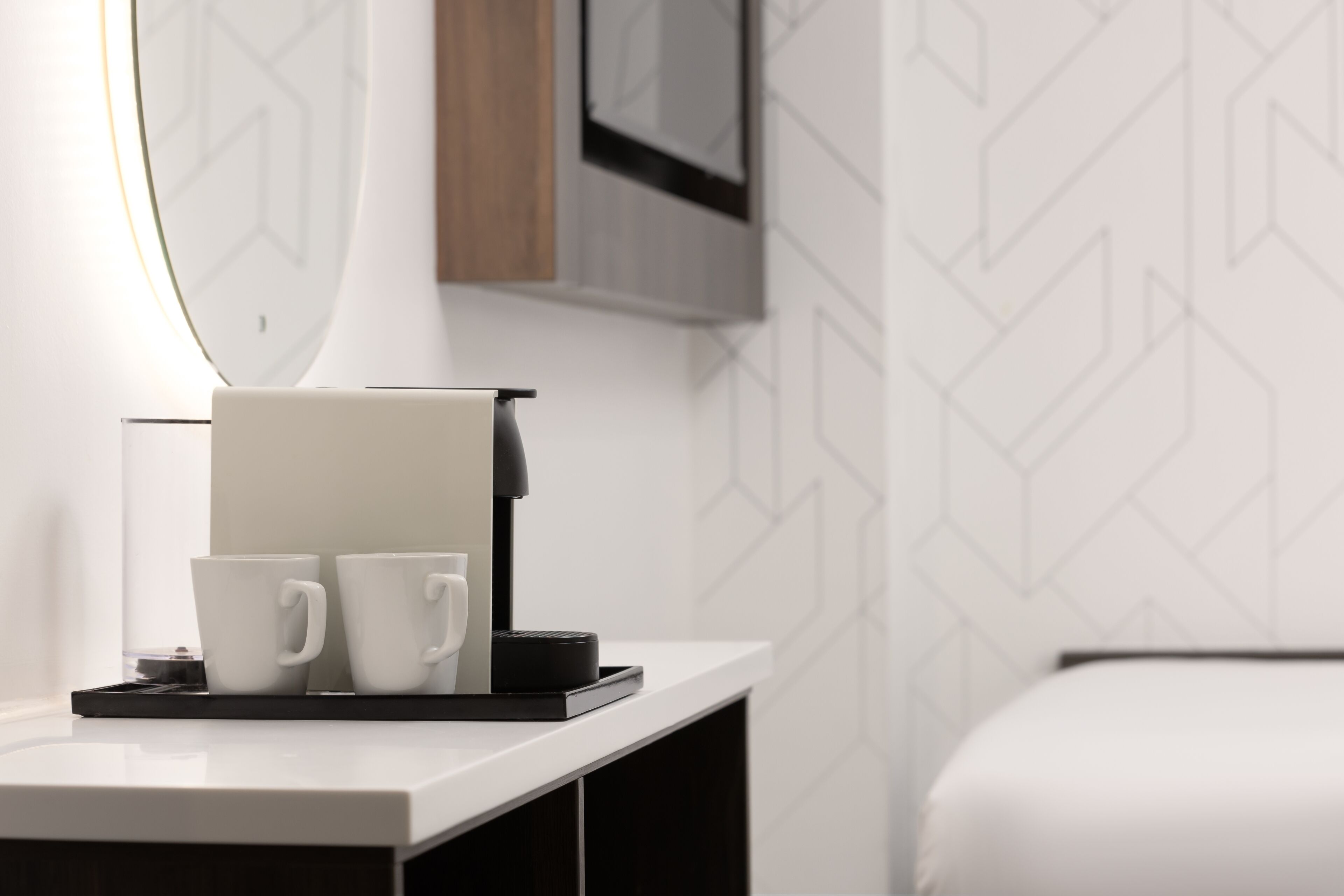 A modern hotel room with a white bed, a bedside table with a coffee machine, and two white cups.