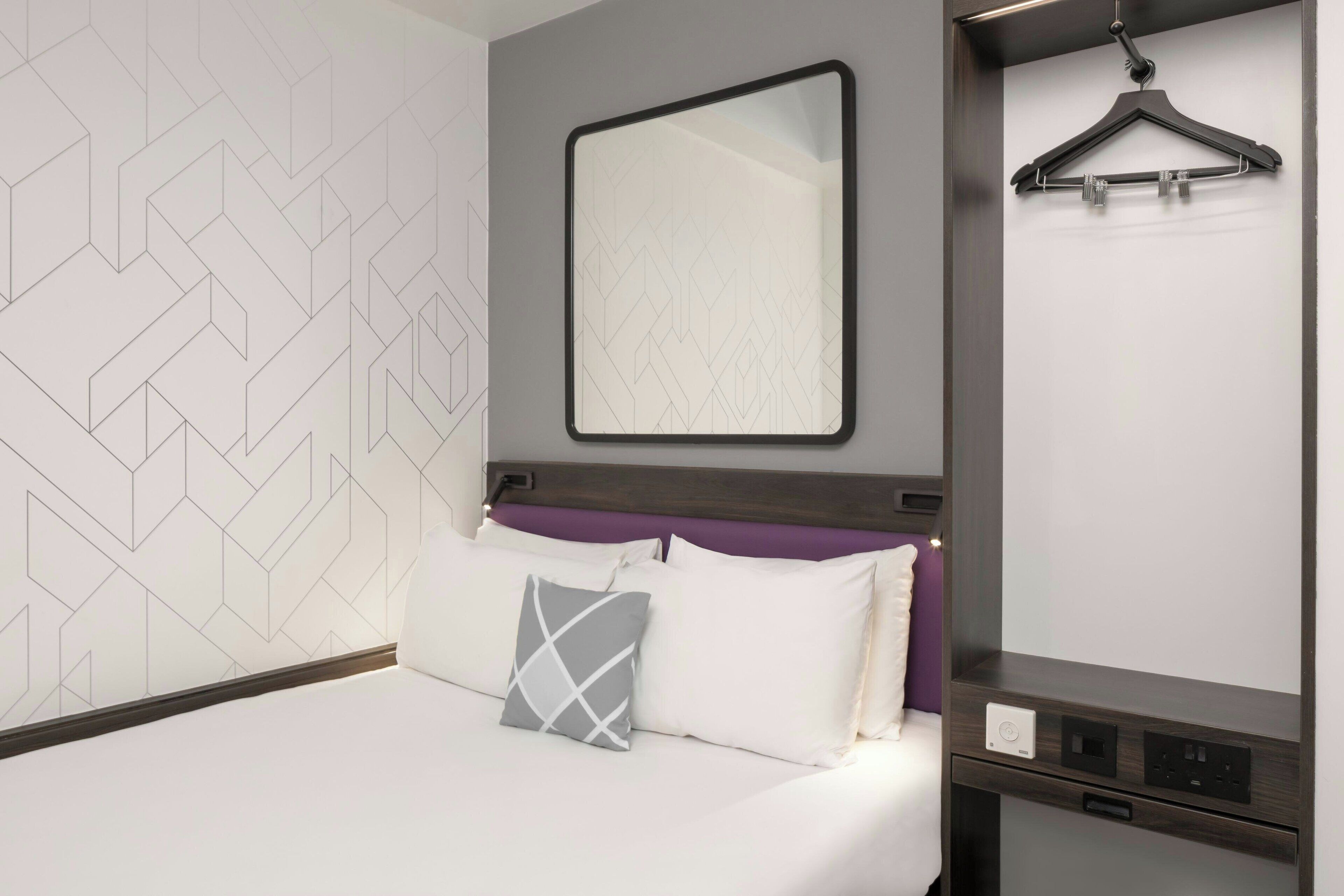 A modern hotel room with a geometric-patterned wall, a bed with white and gray bedding, a purple headboard, a large mirror, and a hanging garment rack.