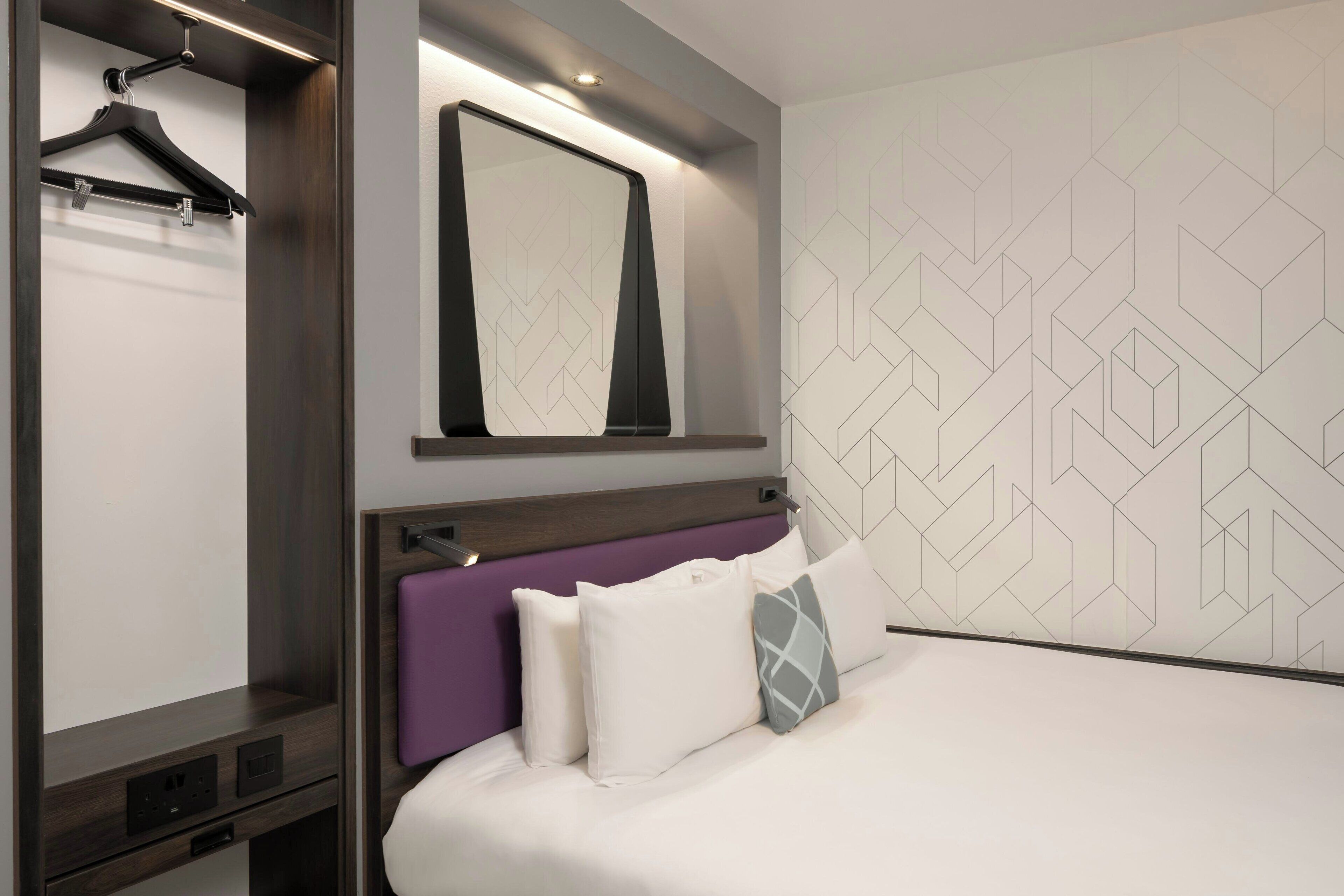 A modern hotel room with a bed, a mirror, a wardrobe, and a geometric patterned wall.