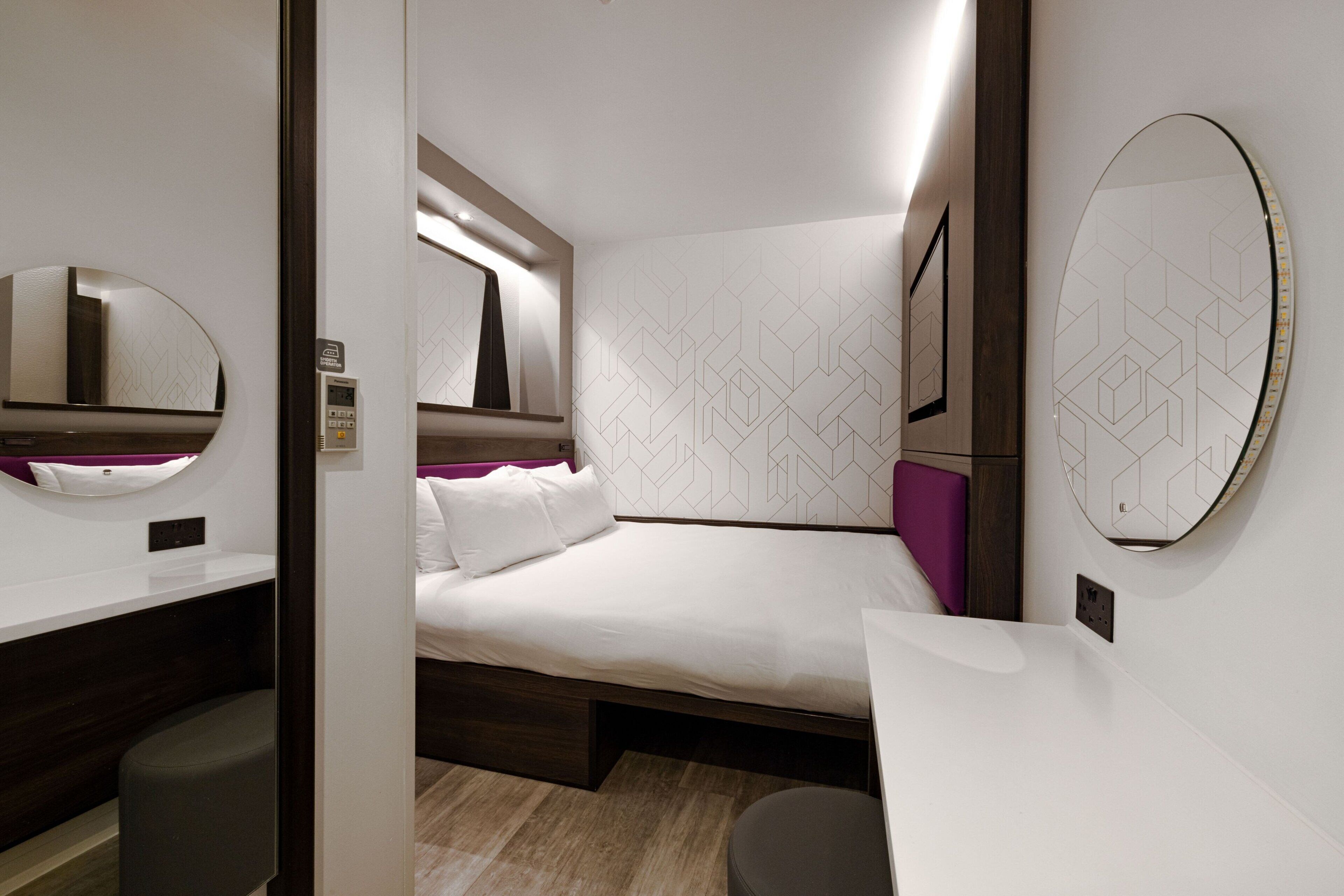 A modern hotel room with a neatly made bed, a bedside table, a chair, and a large mirror.