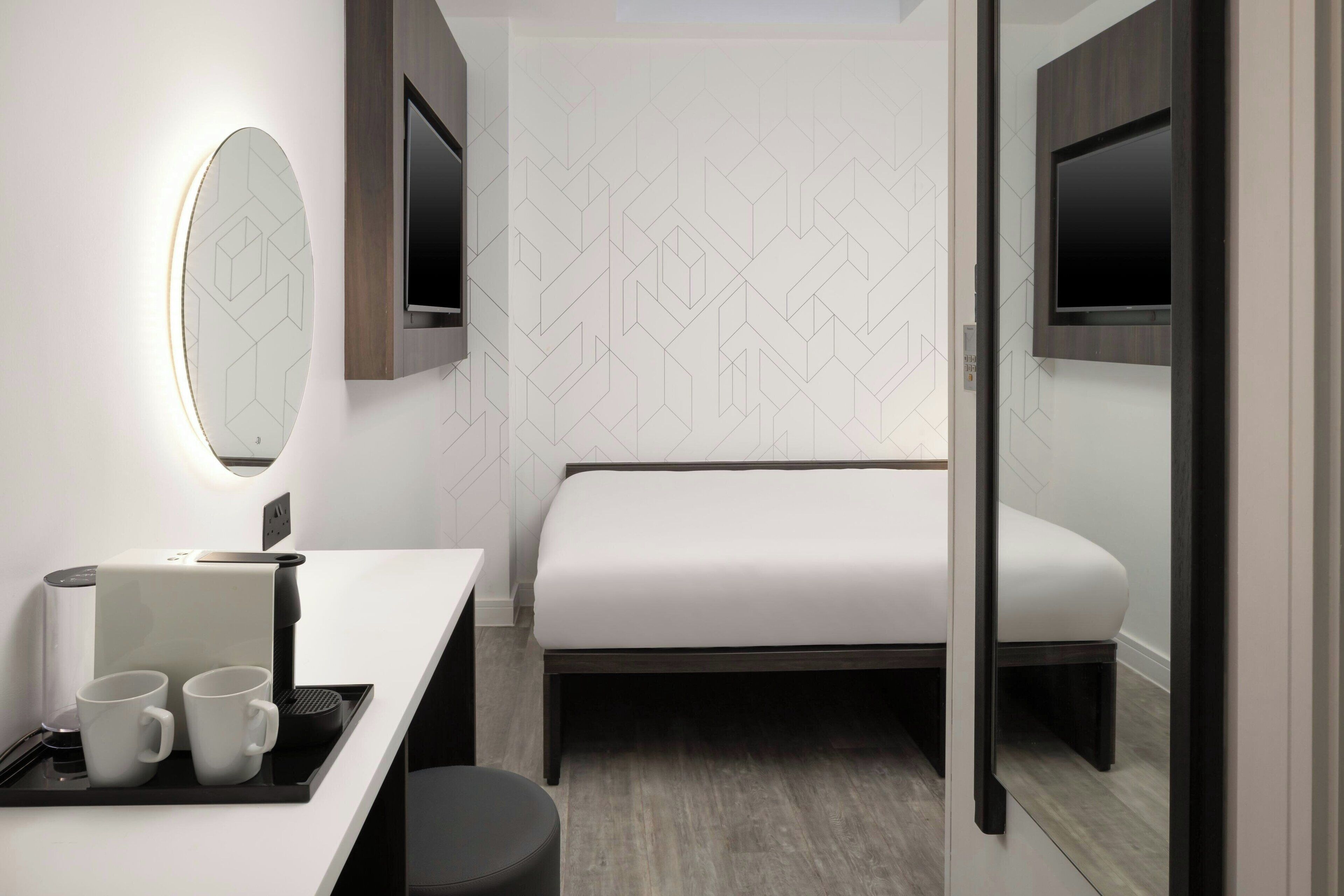 A modern hotel room with a bed, a desk with a coffee maker, two mugs, a mirror, and a TV mounted on the wall.