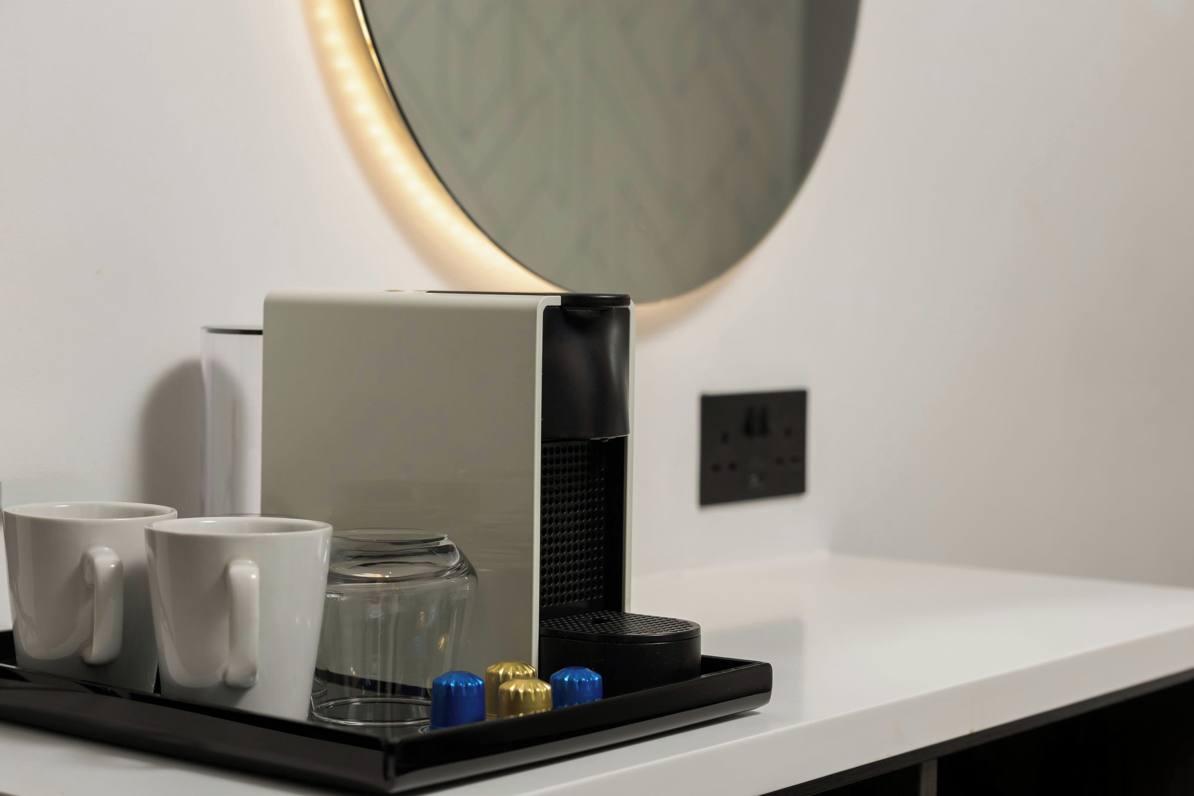 Una cafetera sobre una encimera blanca, con dos tazas blancas y un recipiente de vidrio.