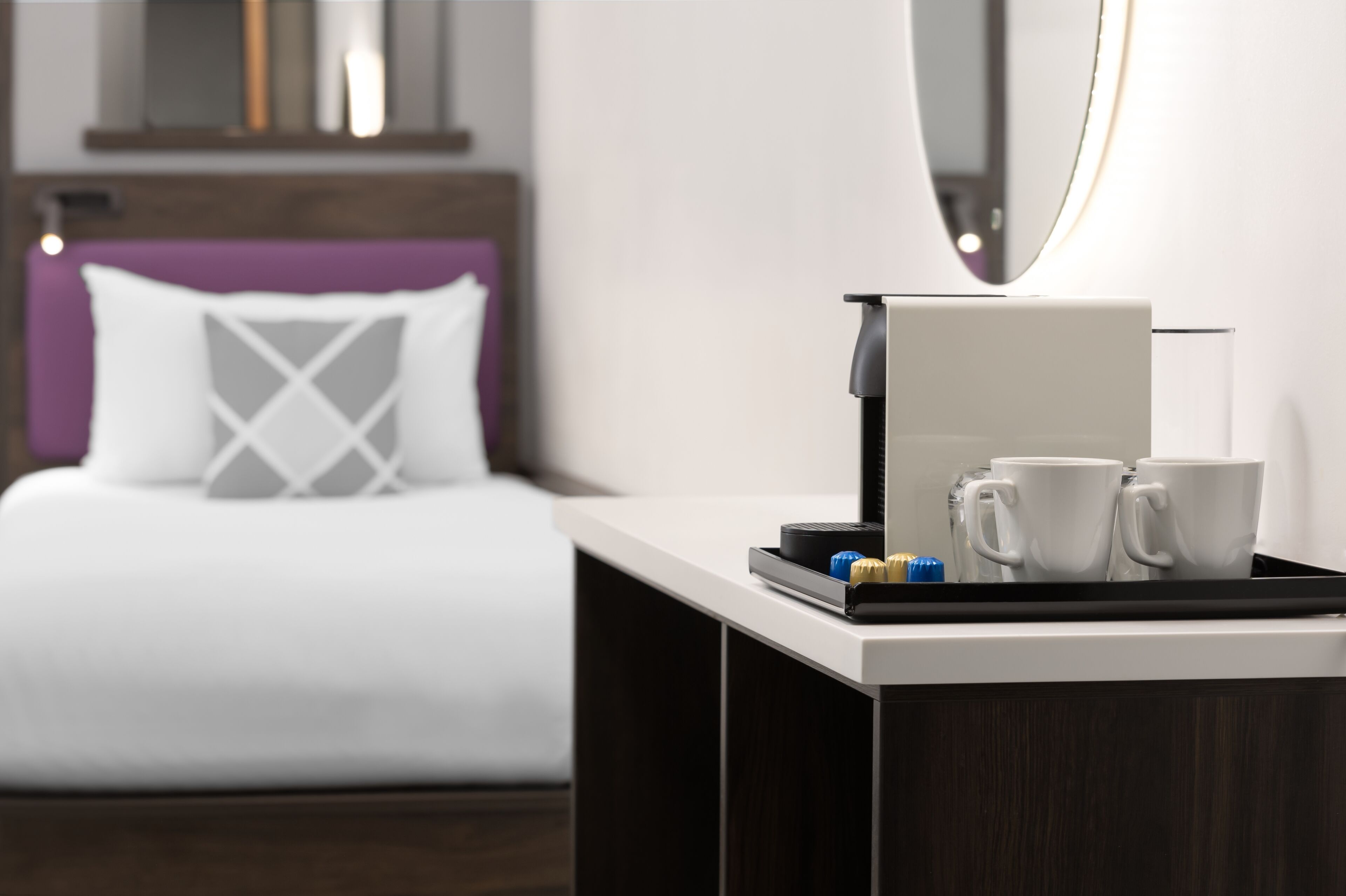 Hotelkamer met een bed, nachtkastje met koffiezetapparaat en twee mokken, en een spiegel erboven.