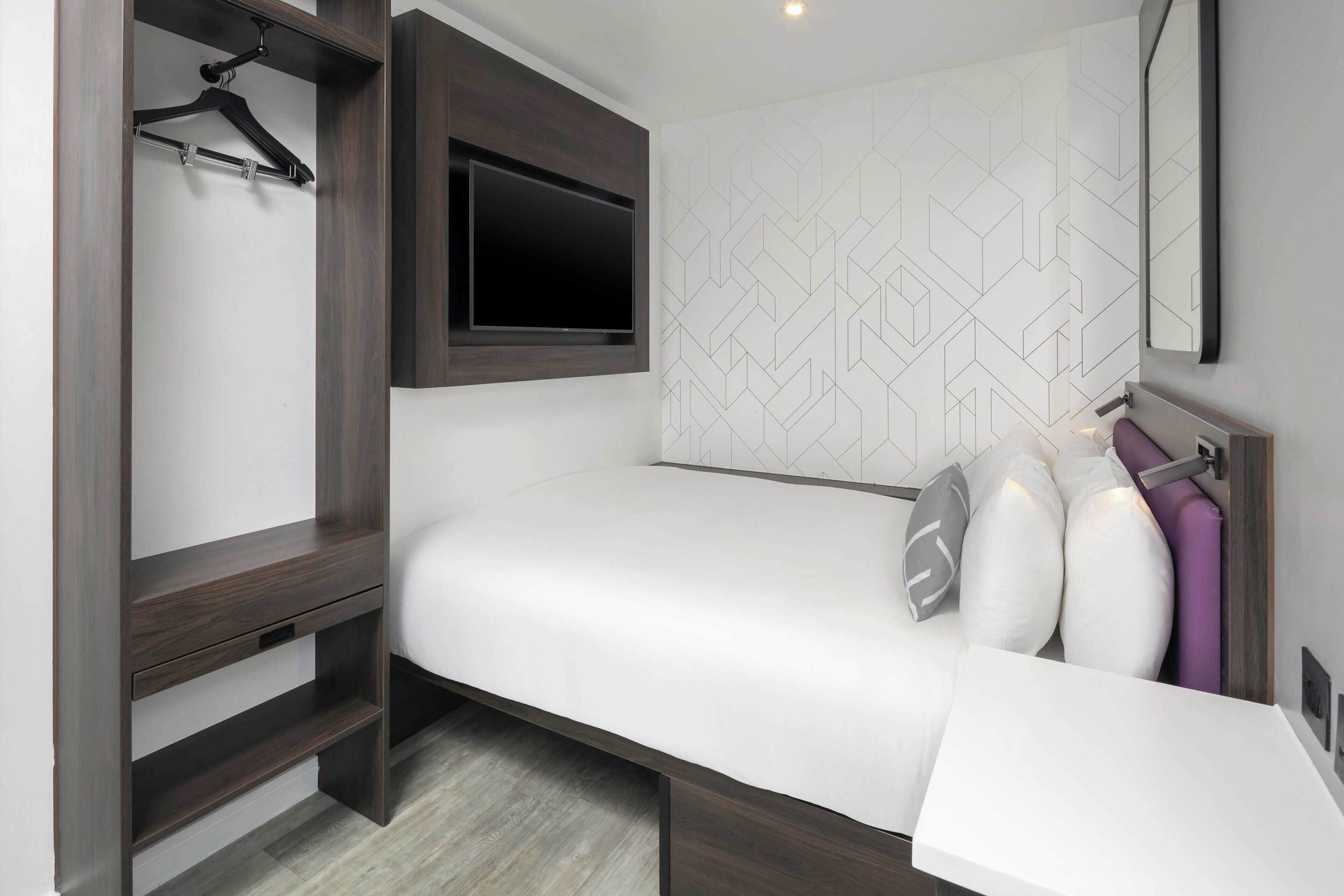 Habitación pequeña con cama, televisor montado en la pared, armario y espejo.