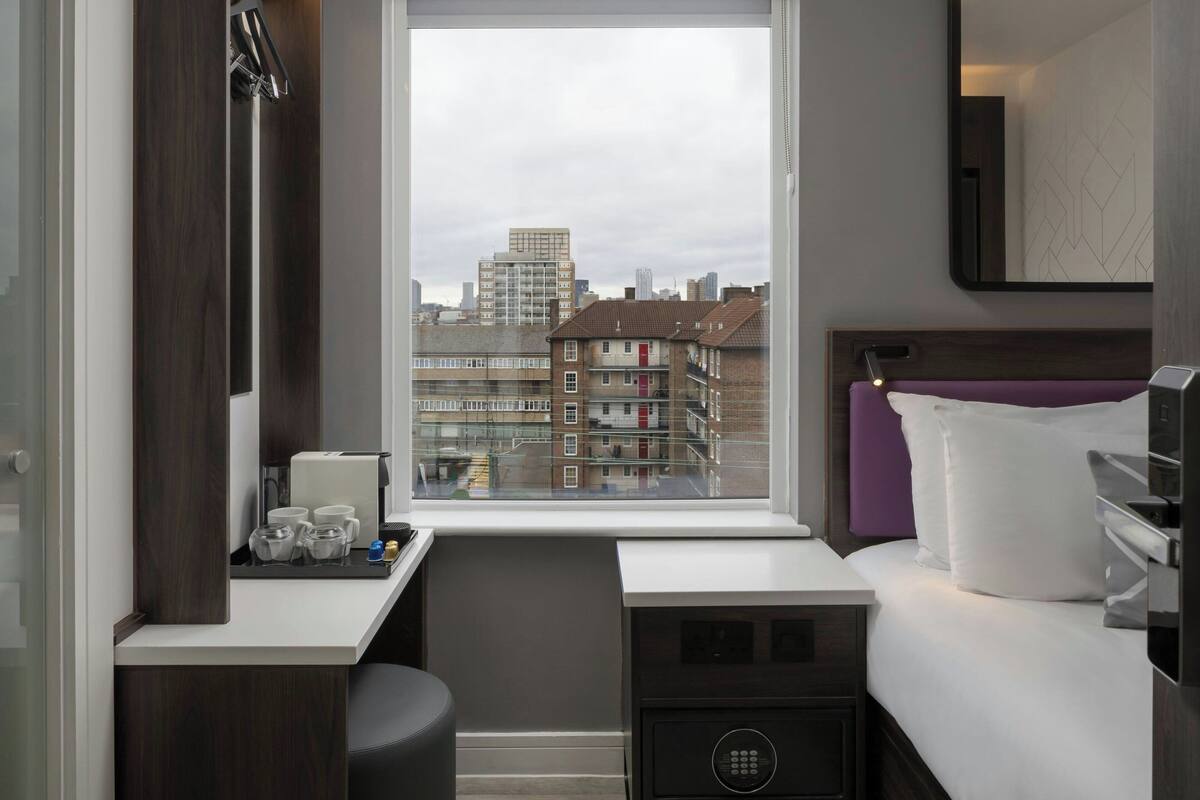 Habitación de hotel con cama, escritorio, silla, ventana con vista a la ciudad y una mesita con objetos encima.