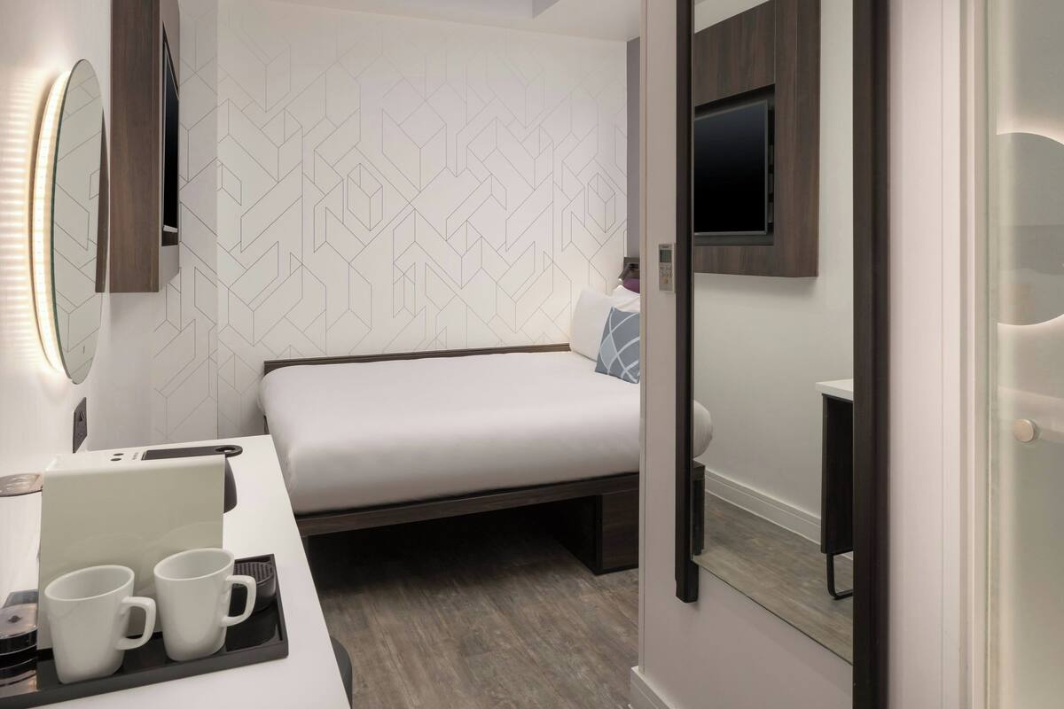 Una habitación de hotel moderna con una cama, una mesita de noche con una computadora portátil y dos tazas, un televisor montado en la pared y un papel tapiz con estampado geométrico.