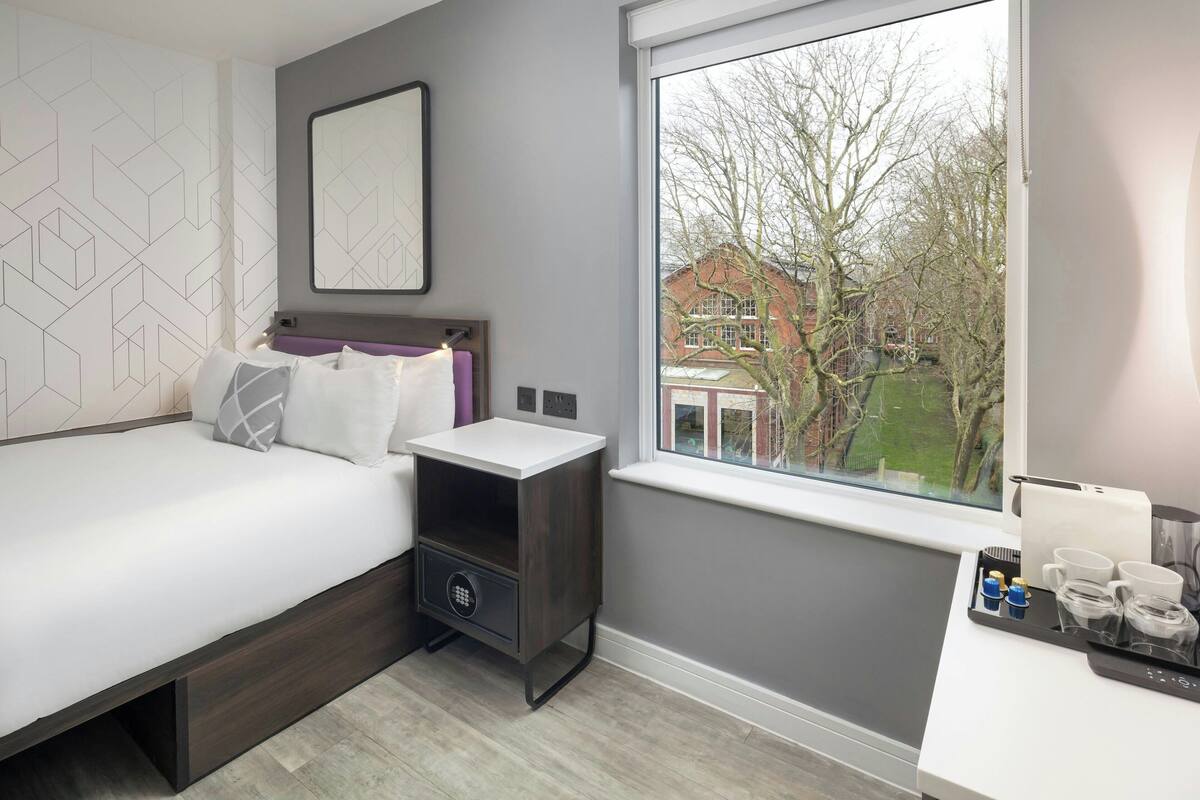 Un dormitorio con cama, mesita de noche y ventana que da a vistas de árboles y edificios.