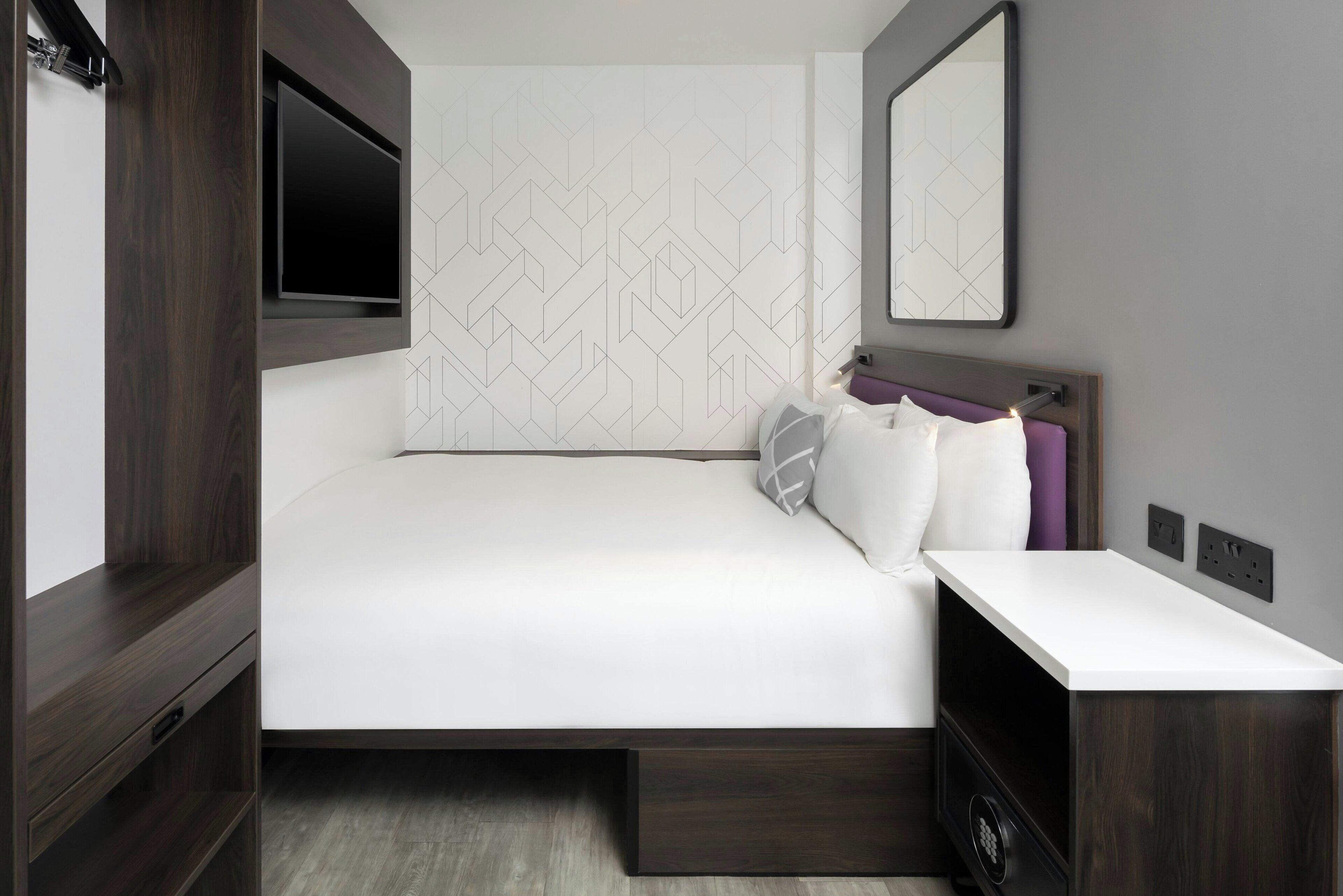 A modern hotel room with a large bed, a bedside table, a TV mounted on the wall, and a mirror above the bed.
