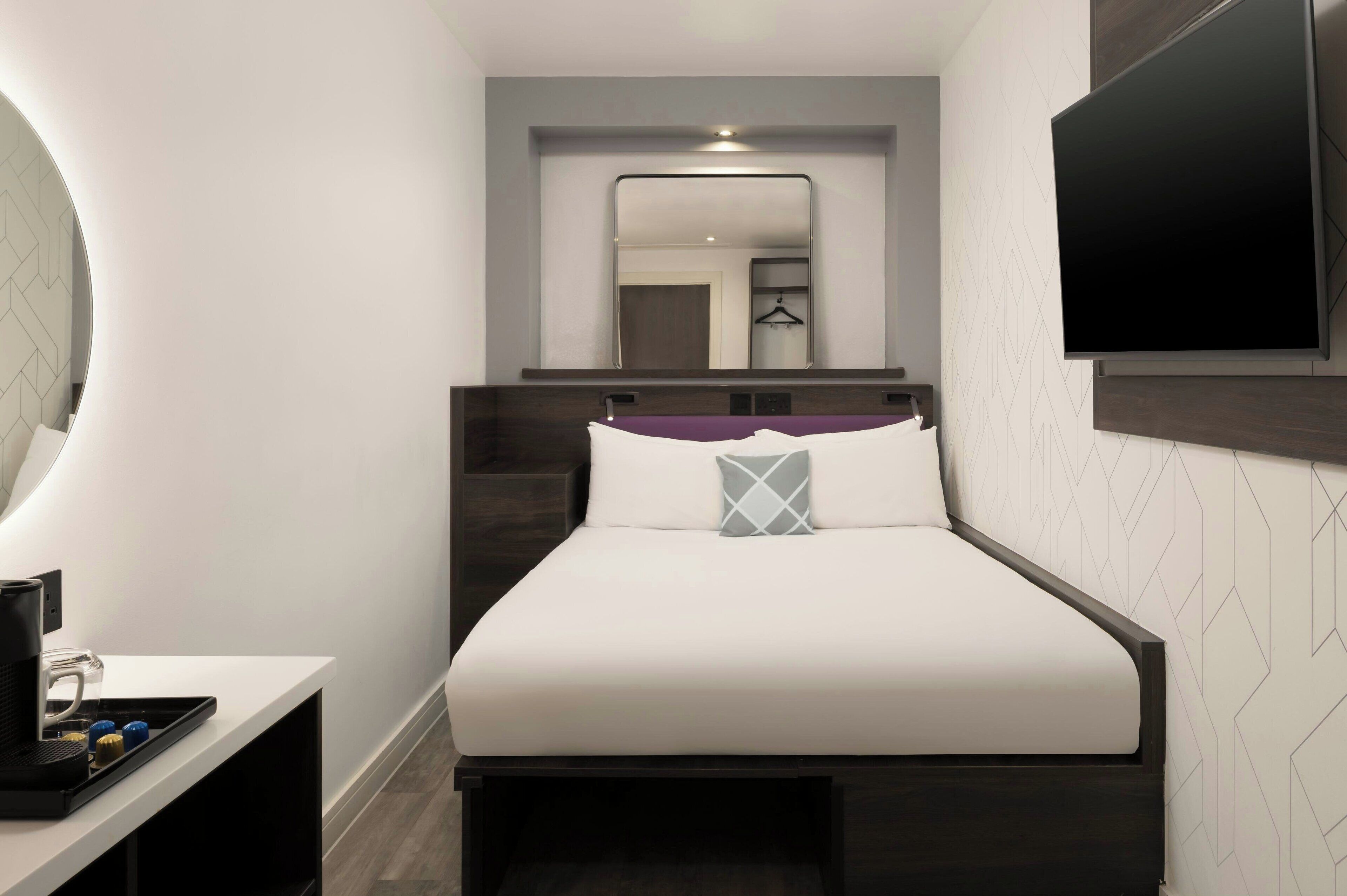 Quarto de hotel moderno com uma cama bem arrumada, uma televisão de tela plana na parede e uma pequena escrivaninha com cafeteira.