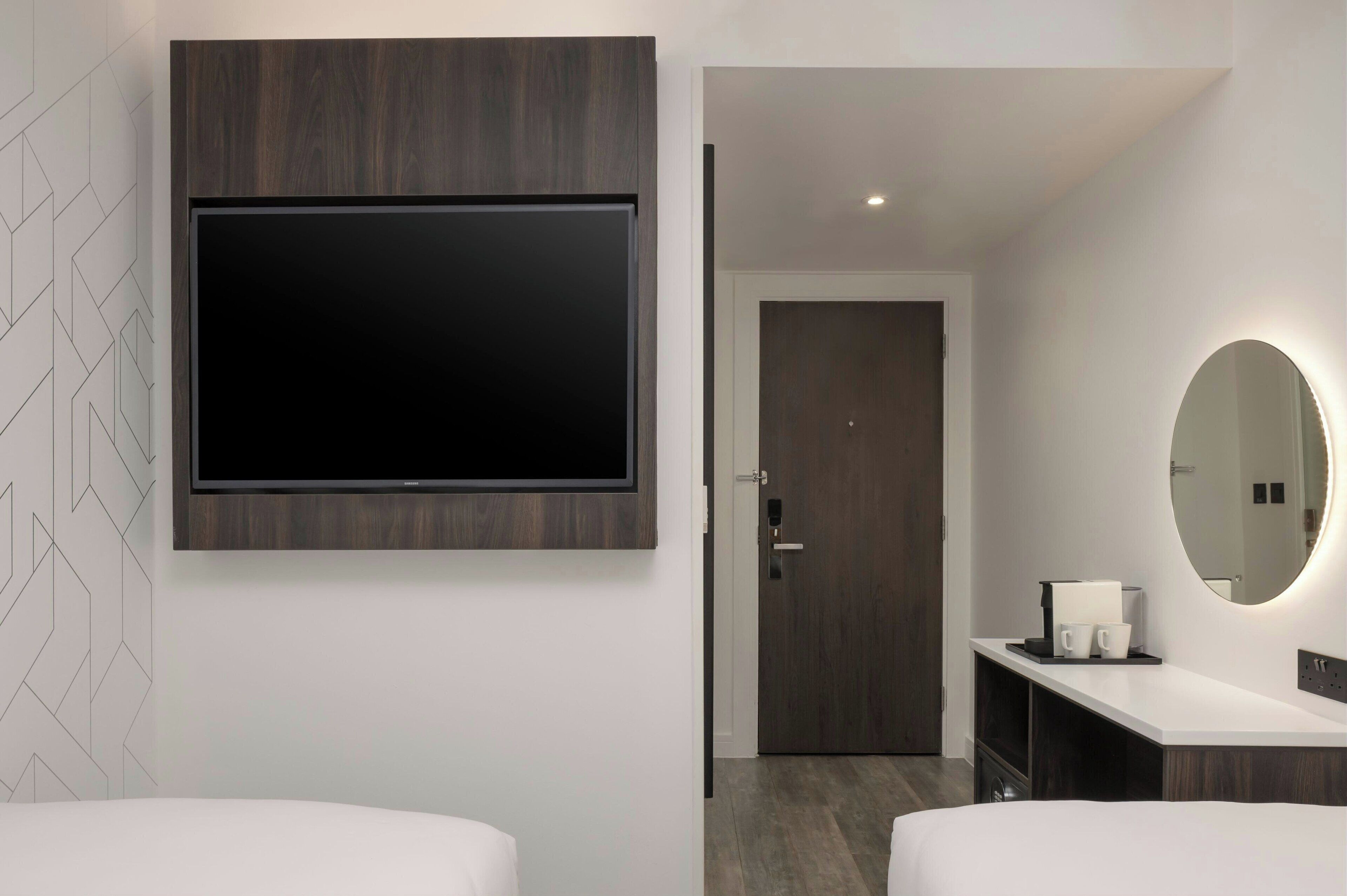 Habitación de hotel moderna con un televisor de pantalla plana grande montado en un panel de madera, un closet amplio con puerta, un espejo redondo con luz y una encimera blanca con cafetera y tazas.
