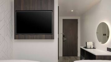 Habitación de hotel moderna con un televisor de pantalla plana grande montado en un panel de madera, un closet amplio con puerta, un espejo redondo con luz y una encimera blanca con cafetera y tazas.