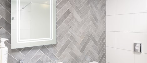 A modern bathroom with a herringbone tile backsplash, a white sink, a mirror with a light, and a toilet.