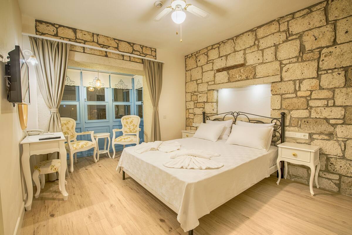 Een slaapkamer met een stenen muur, een bed met witte lakens, een bureau met stoel en een televisie aan de muur.