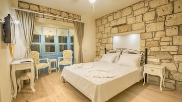 A bedroom with a stone wall, a bed with white linens, a desk with a chair, and a television on the wall.