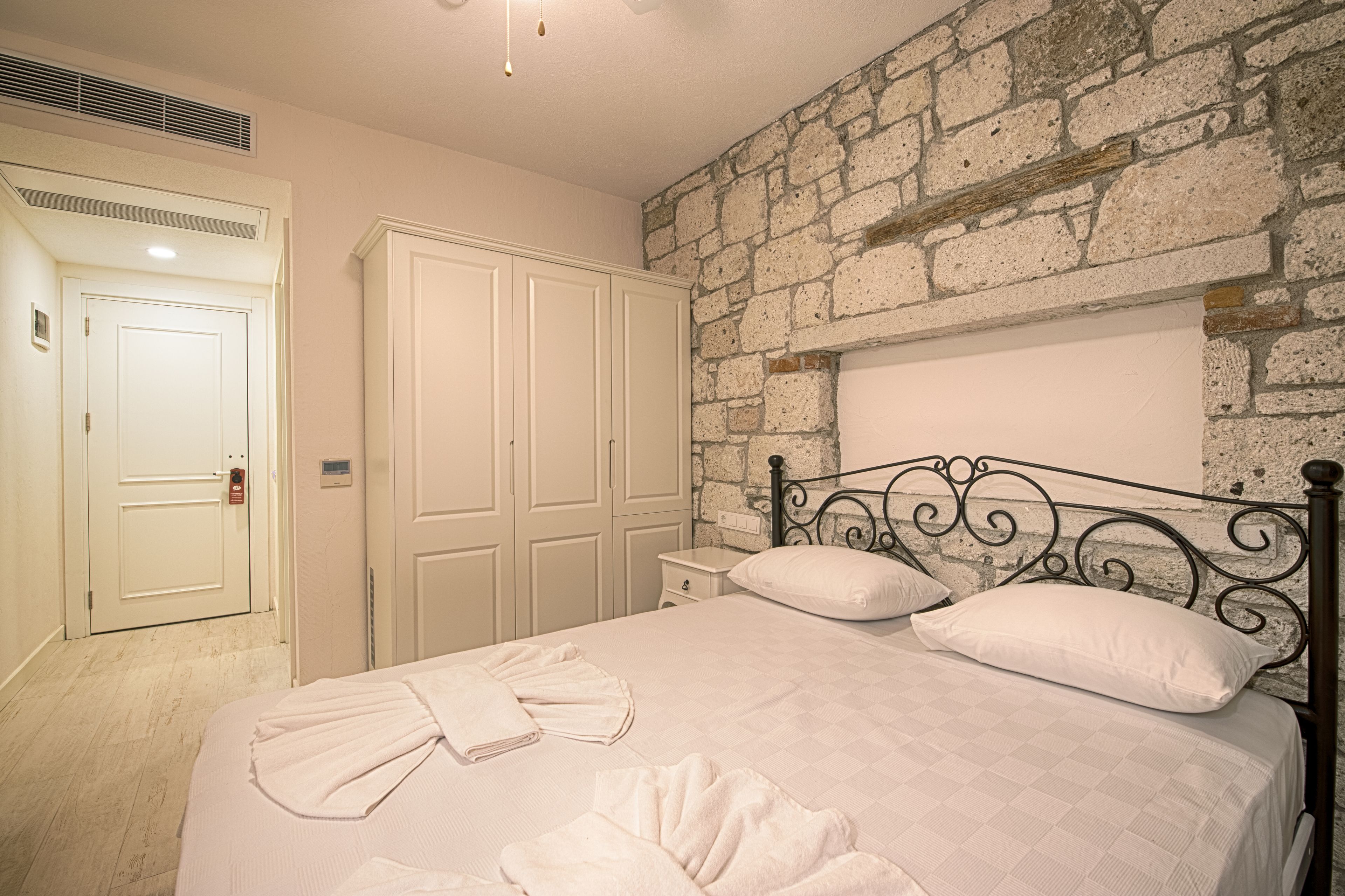 A bedroom with a stone wall, a bed with white bedding, a metal headboard, and a white wardrobe.