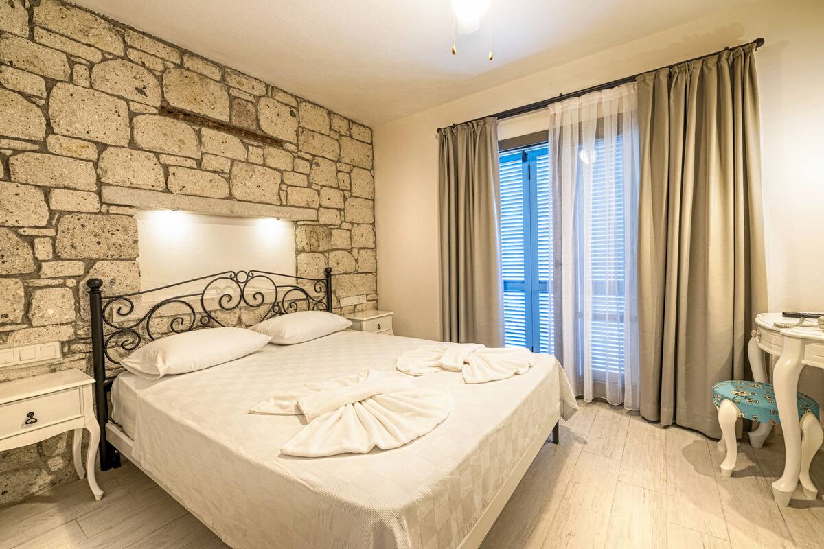 Een net opgemaakt bed met handdoeken, een stenen muur, een nachtkastje en een klein tafeltje met een stoel.