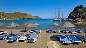 Een strand met ligbedden en parasols, een heldere blauwe zee en boten afgemeerd bij een rotsachtige klif.
