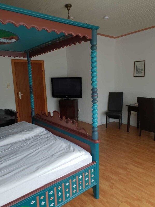 Un lit à baldaquin avec un cadre en bois et un matelas blanc, un téléviseur posé sur un meuble, un bureau et une chaise dans une pièce dont le sol est recouvert de planches de bois.