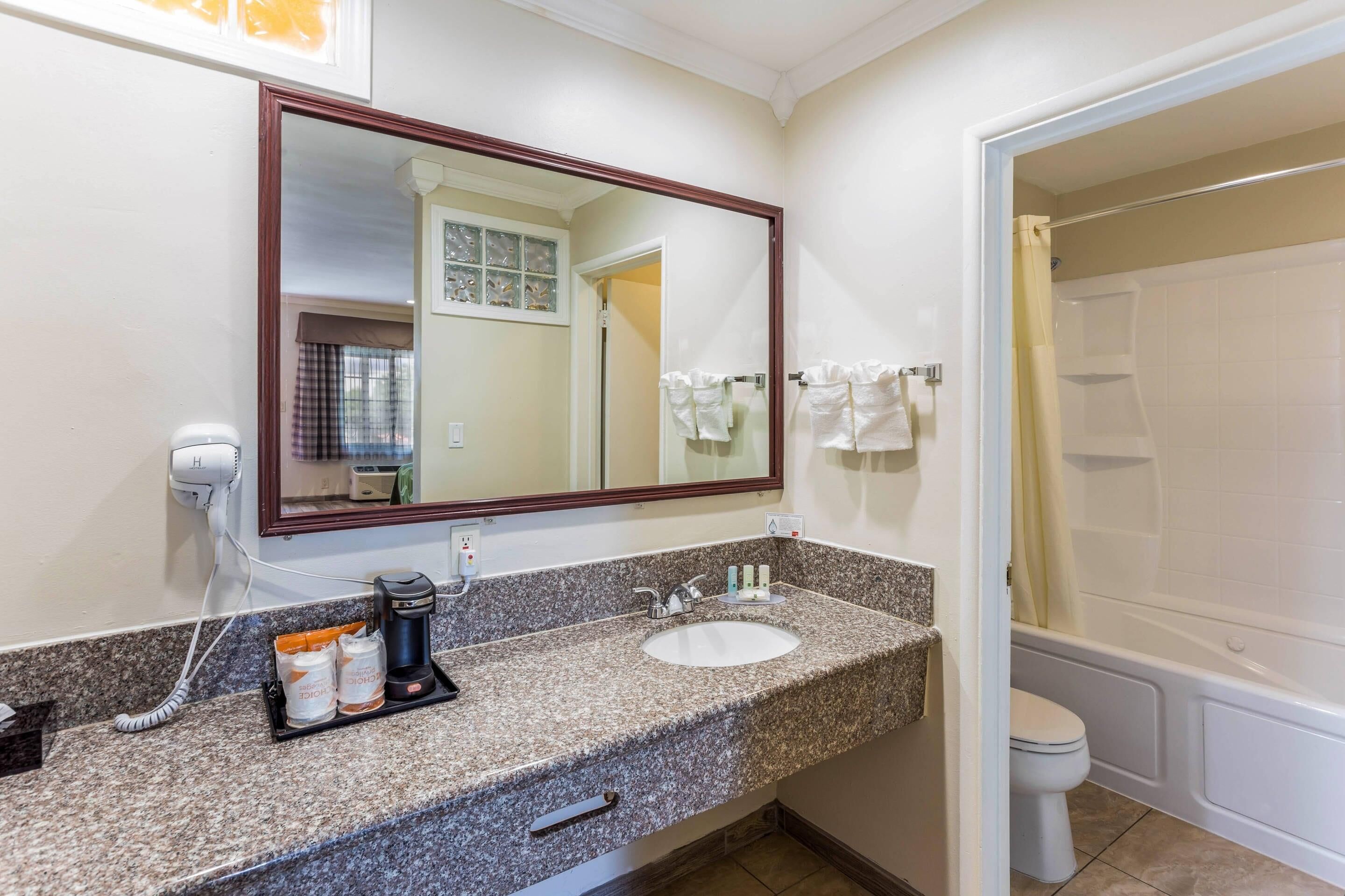 A hotel bathroom with a granite countertop, a sink, a mirror, and a shower area.