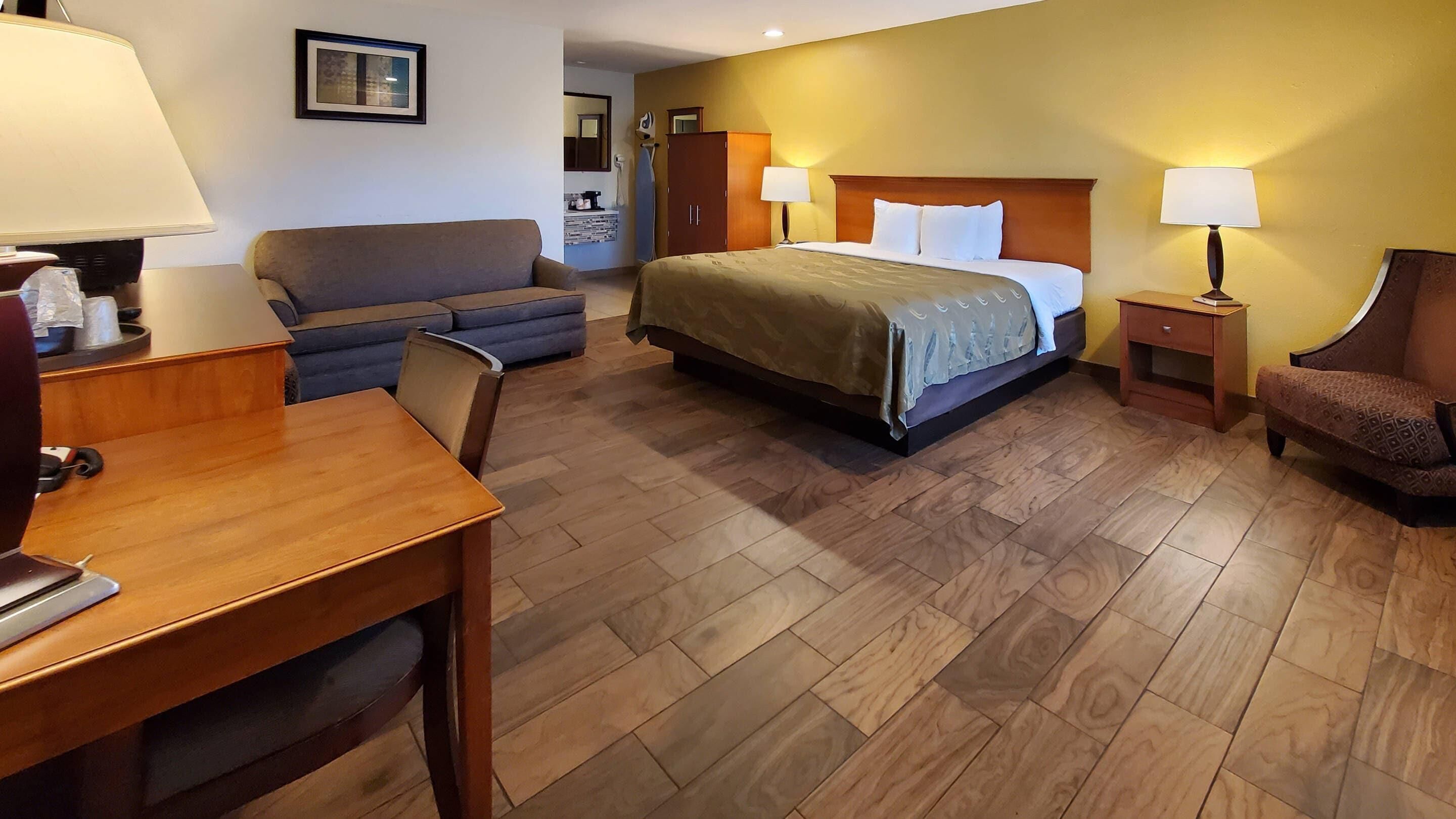 A hotel room with a bed, a desk, a chair, a sofa, and a nightstand with a lamp.