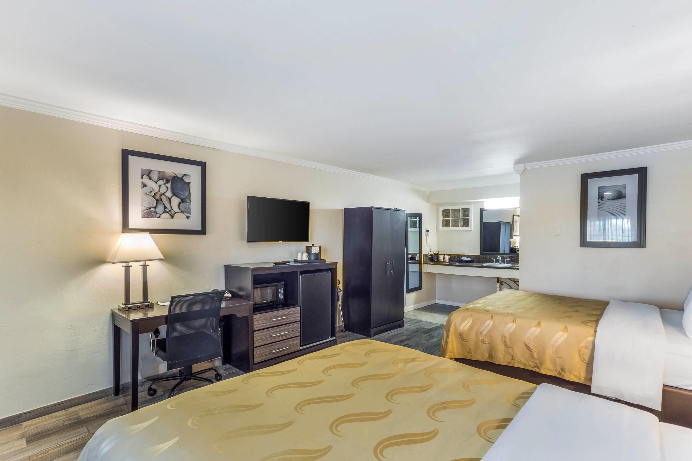 A hotel room with two beds, a desk with a computer, a television, and a kitchenette.